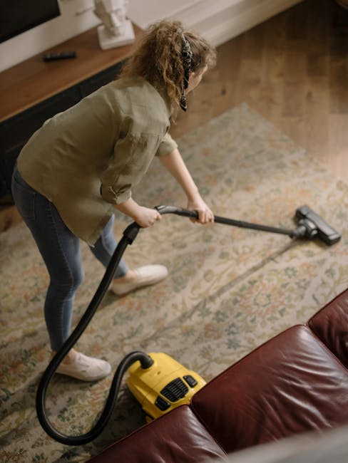 A woman operating a yellow vacuum cleaner with a long hose attachment on a floral-patterned carpet within a living room. She is leaning forward, wearing a beige shirt, dark jeans, and white shoes. The