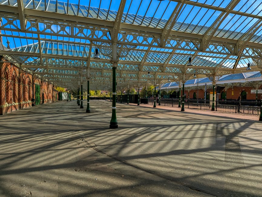 An outdoor covered waiting area at Wood Green station featuring a glass and metal roof with intricate latticework, supported by green and black lamp posts. The paved ground is clean with visible shado