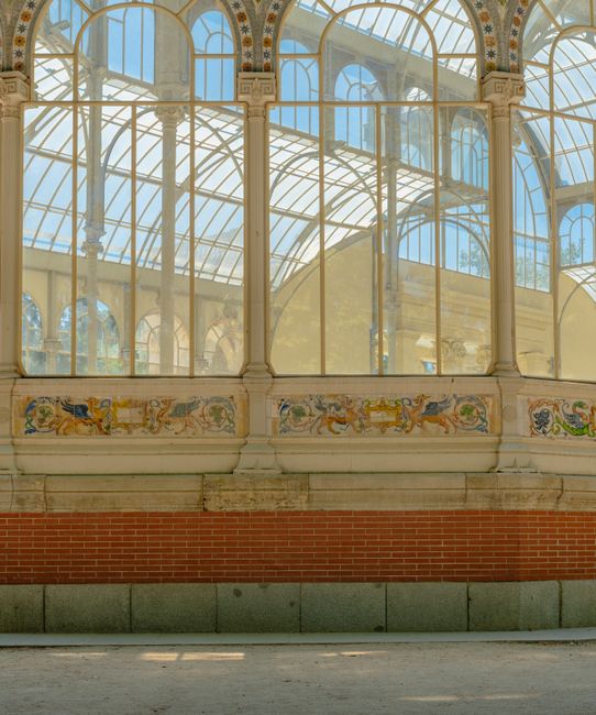An outdoor covered waiting area at Wood Green station featuring a glass and metal roof with intricate latticework, supported by green and black lamp posts. The paved ground is clean with visible shado